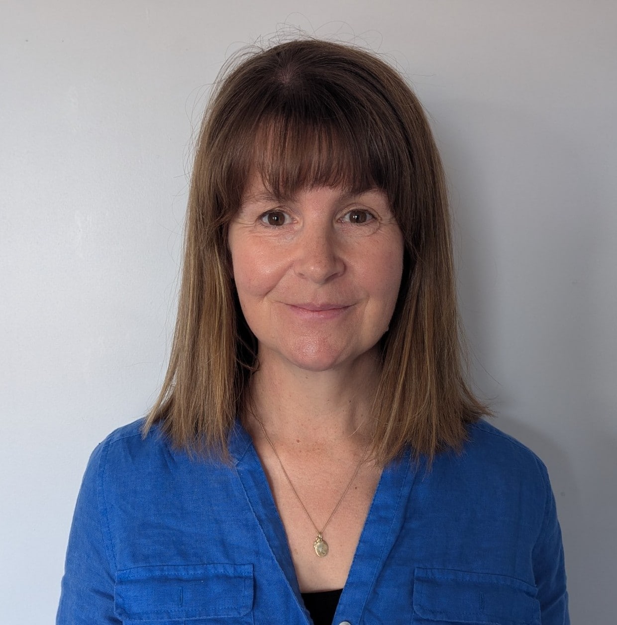 Smiling woman with shoulder-length brown hair and bangs wearing a blue button-up shirt, posed against a light gray wall.