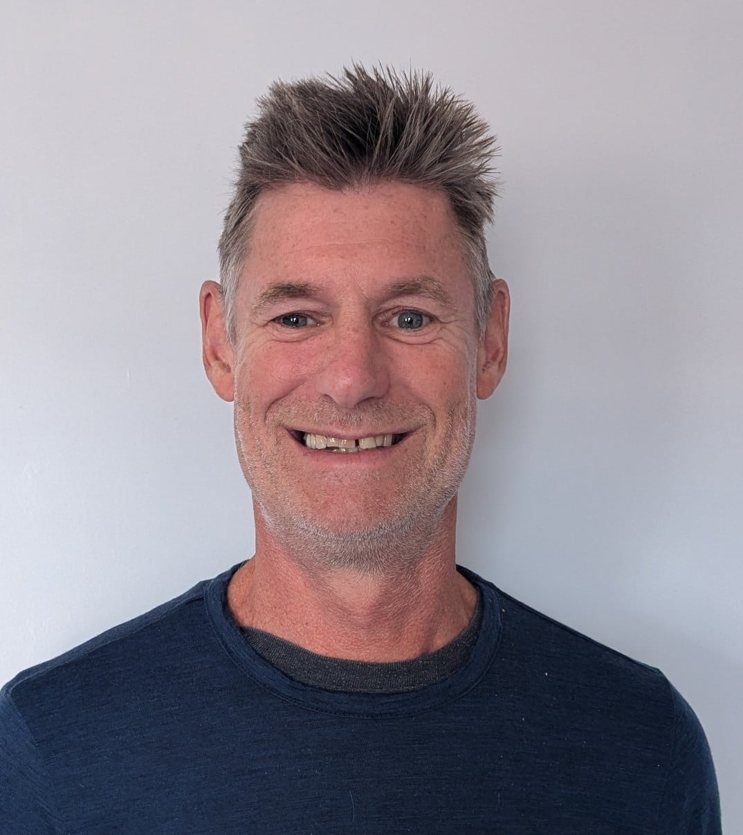 Smiling man with short, spiky salt‑and‑pepper hair wearing a dark blue crewneck shirt, against a light gray wall (head-and-shoulders portrait).